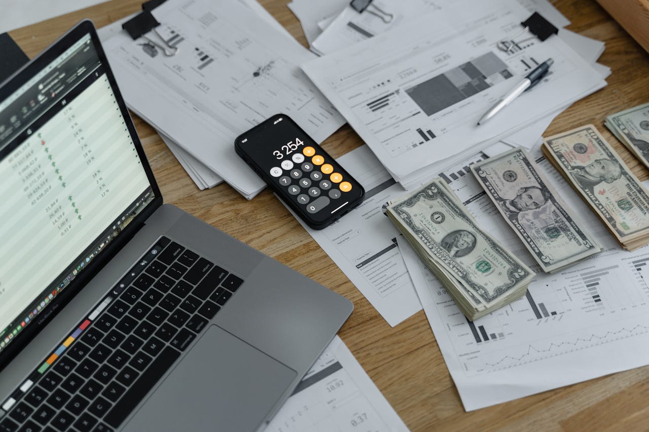 A desk with financial documents, currency, a laptop, and phone calculator.